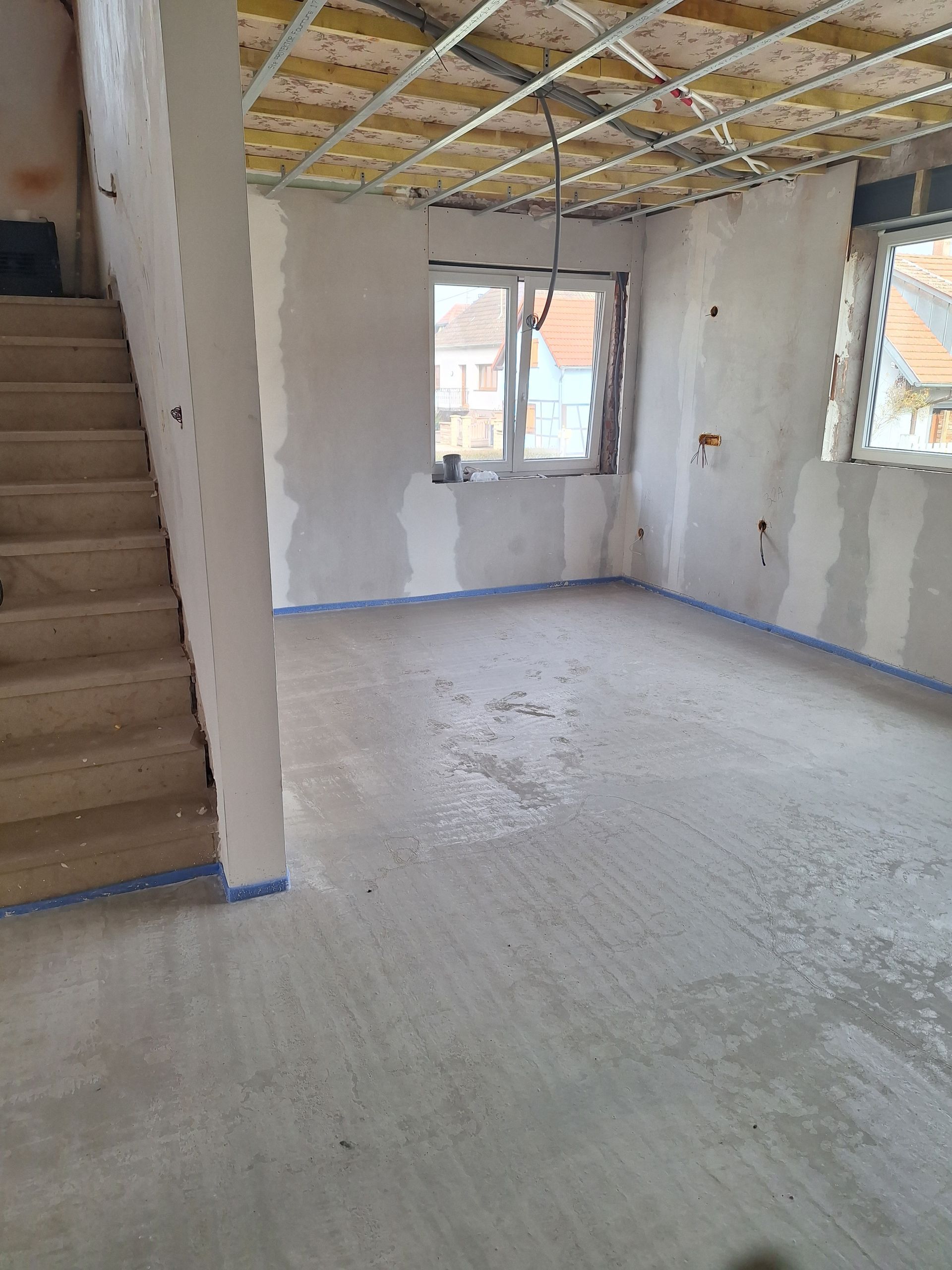 Intérieur en cours d'aménagement avec escalier, fenêtres, sol en béton et charpente apparente.