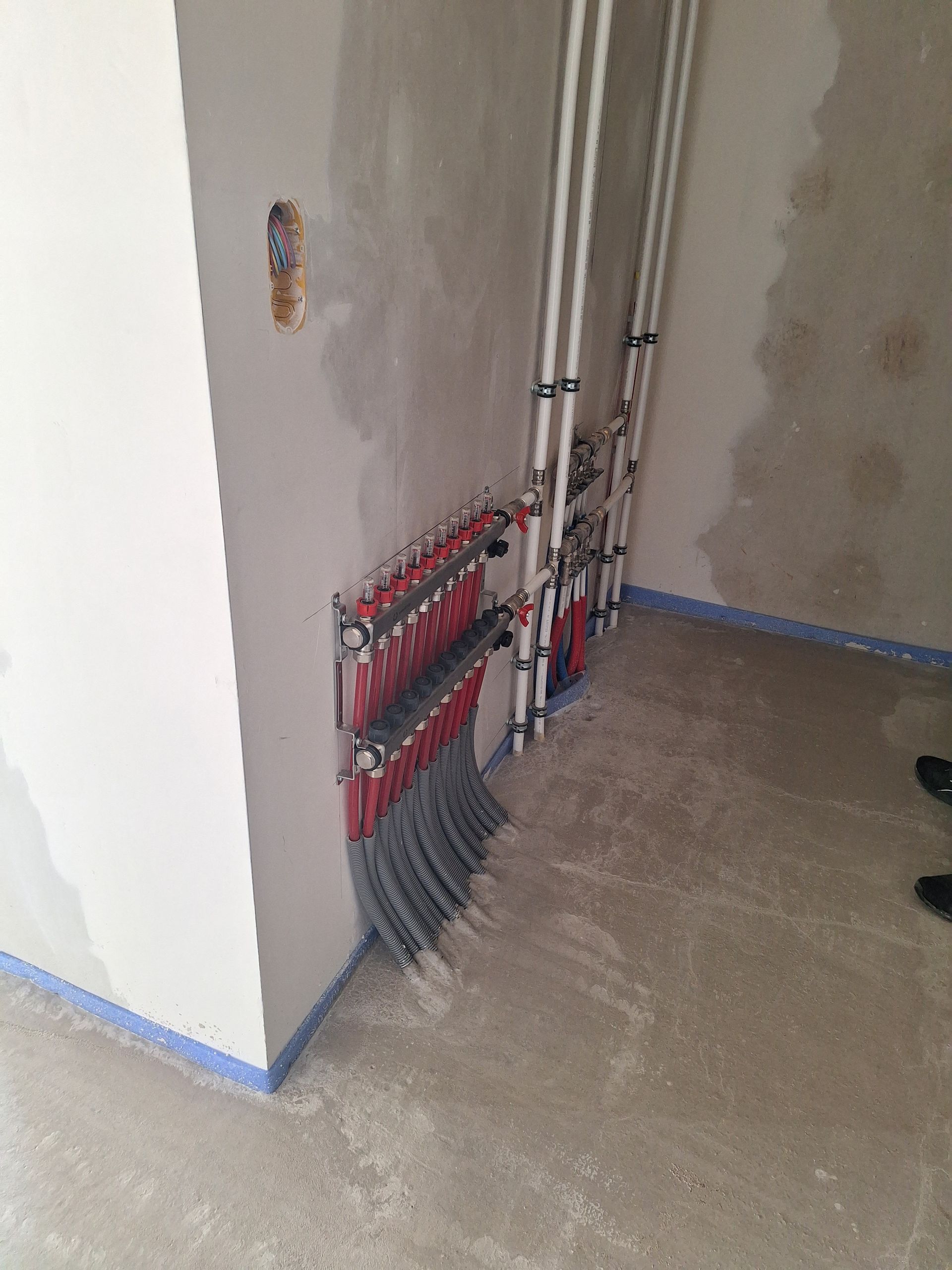 Tuyauterie et vannes installées dans un coin d'une pièce en construction. Sol en béton gris, murs blancs.