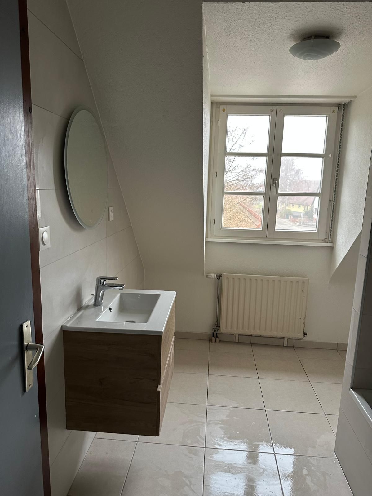 Salle de bains avec murs blancs, fenêtre, meuble-lavabo en bois, lavabo et miroir rond.