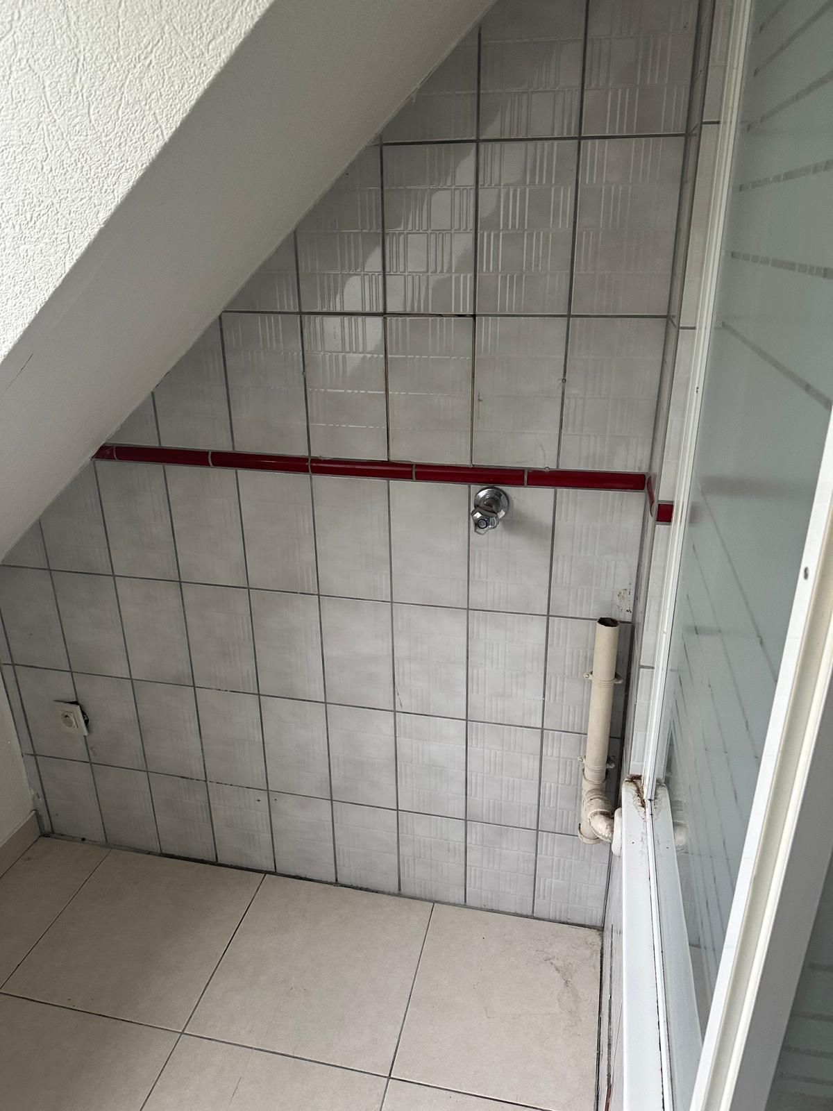 Salle de bains avec plafond en pente, murs et sol carrelés, douche et porte partiellement visible.