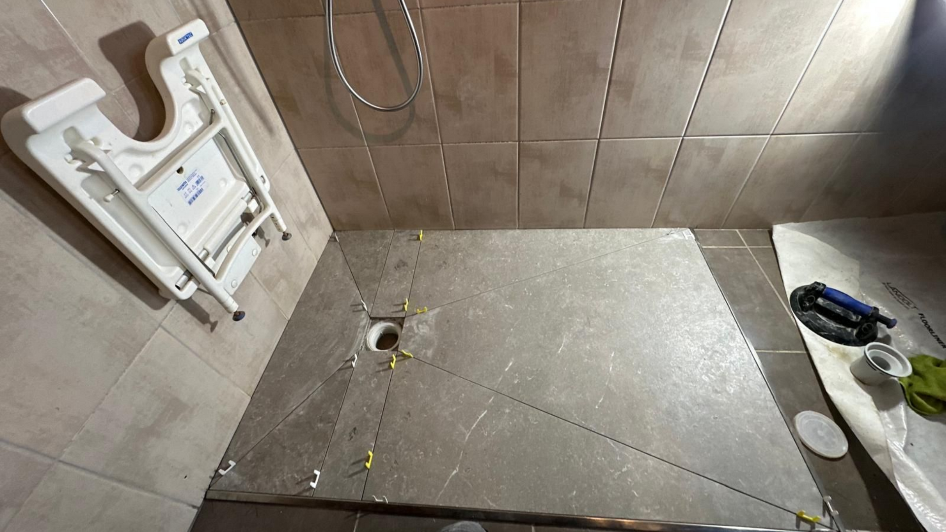 Cabine de douche avec murs et sol carrelés, bonde centrale. Un siège rabattable mural est présent.