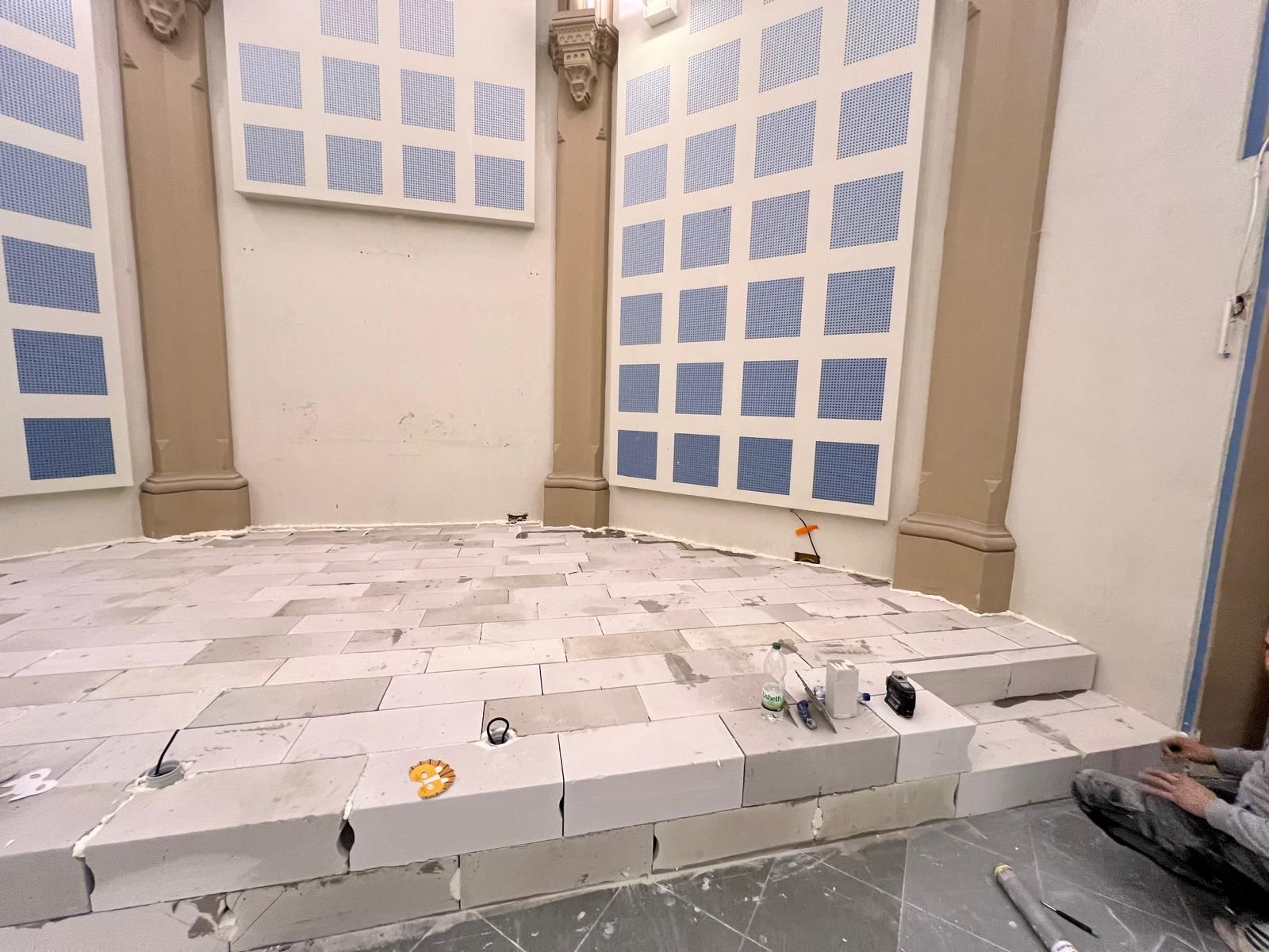 Chantier : un ouvrier pose des blocs gris clair sur un sol incurvé dans une pièce aux murs percés de fenêtres bleues et aux piliers beiges.