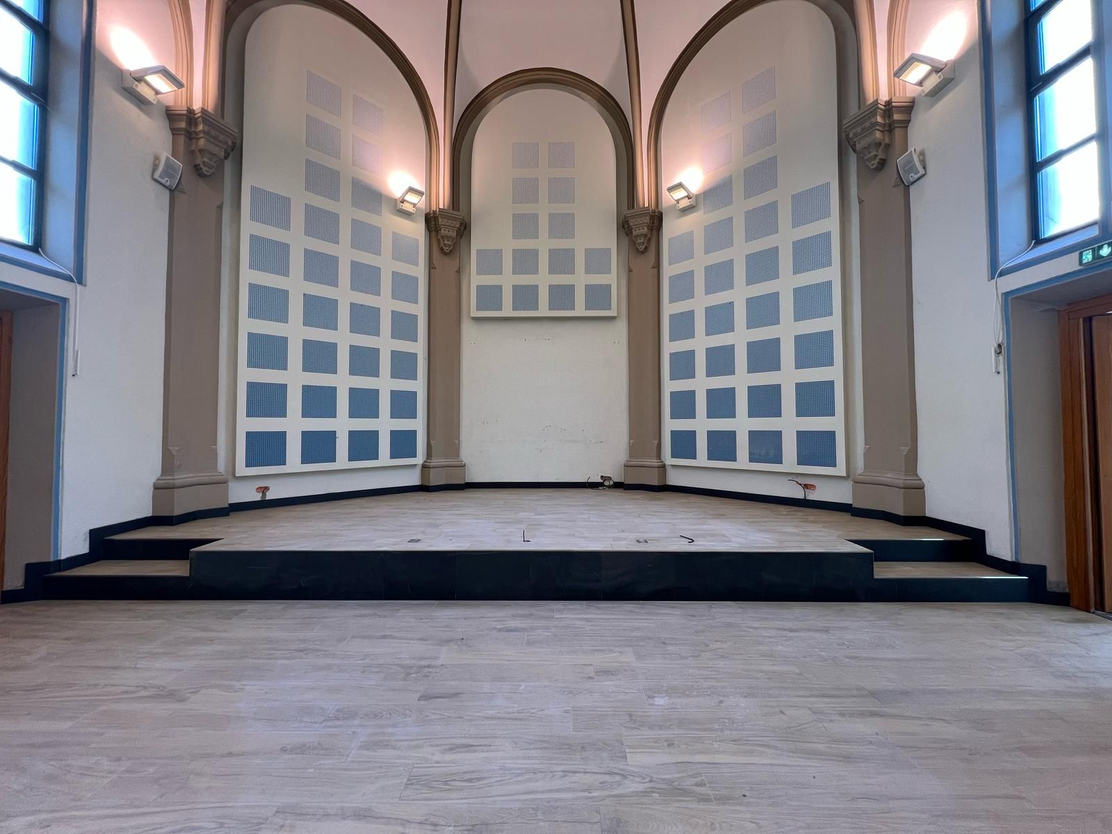 Une scène vide dans une pièce aux murs blancs, aux alcôves cintrées et aux panneaux muraux acoustiques bleus et blancs.