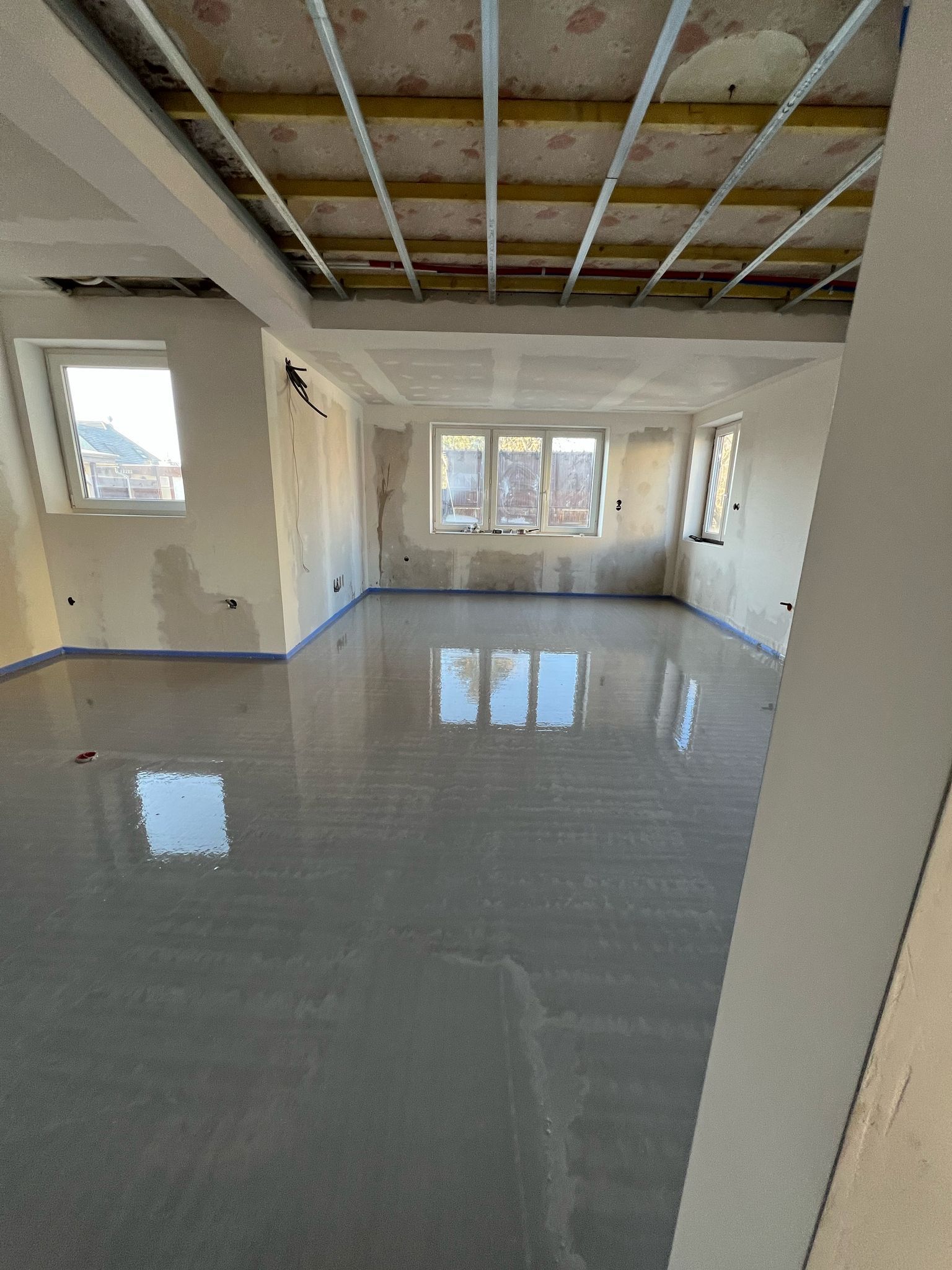 Pièce intérieure en cours de construction avec nouveau sol en ciment, fenêtres et charpente de plafond apparente.