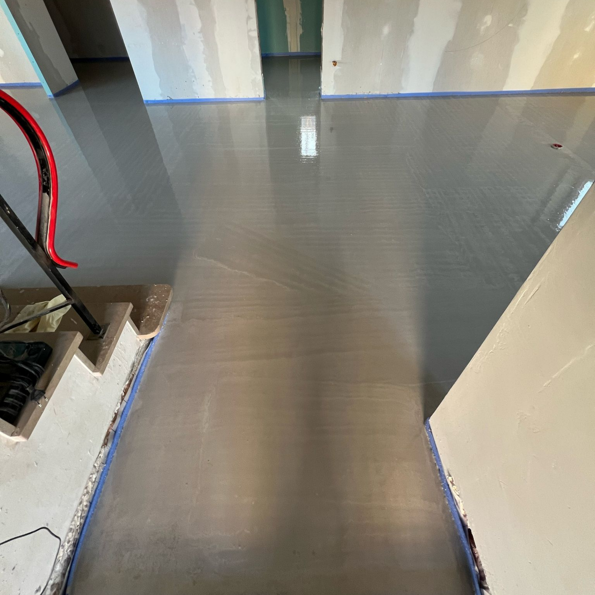 Sol en béton gris fraîchement coulé dans une pièce en construction, avec du ruban adhésif bleu le long des murs et un tuyau rouge.