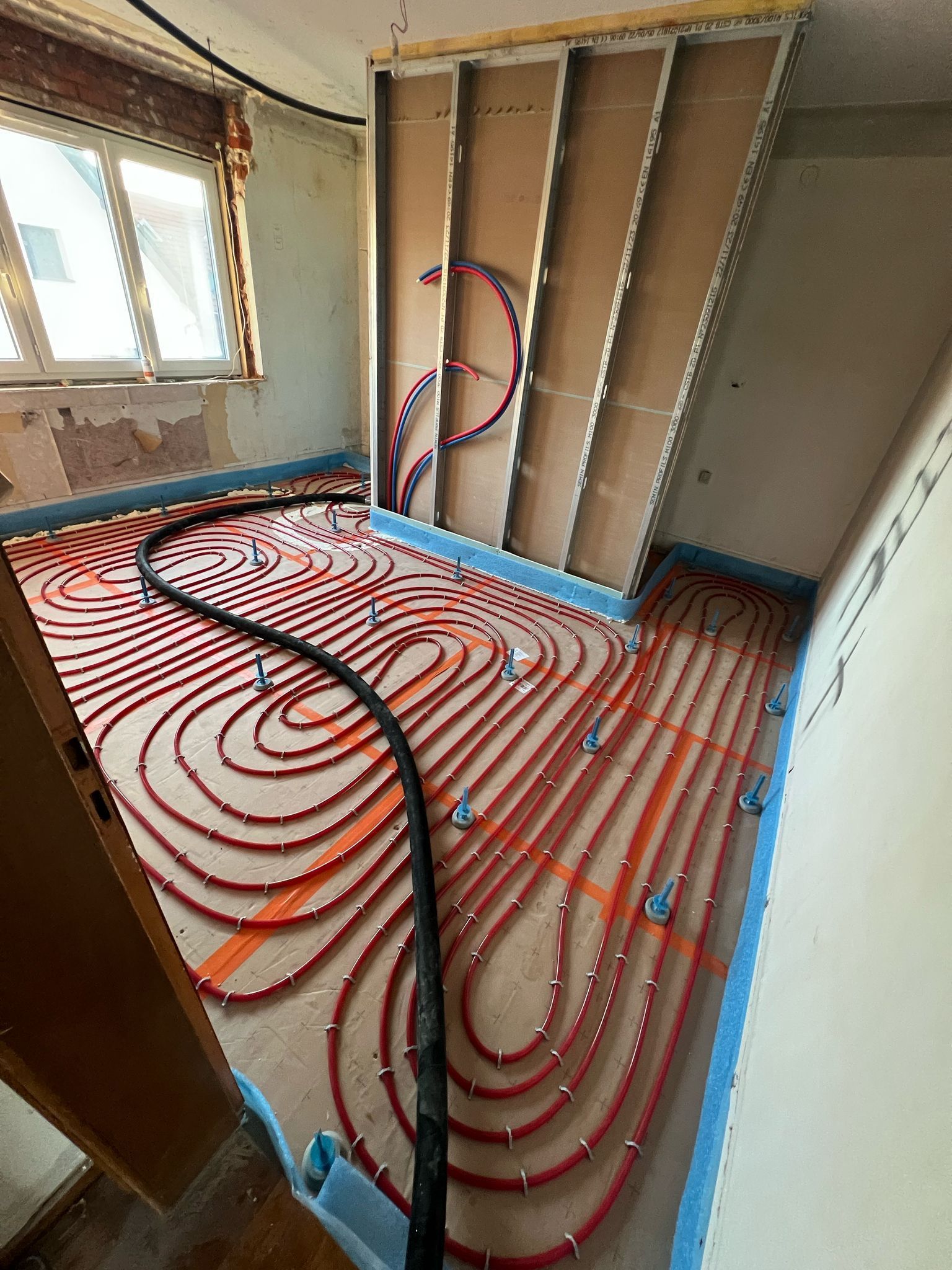 Des tubes de chauffage au sol radiant rouges ont été posés sur un sol en construction ; pièce avec fenêtre et cloisons sèches.