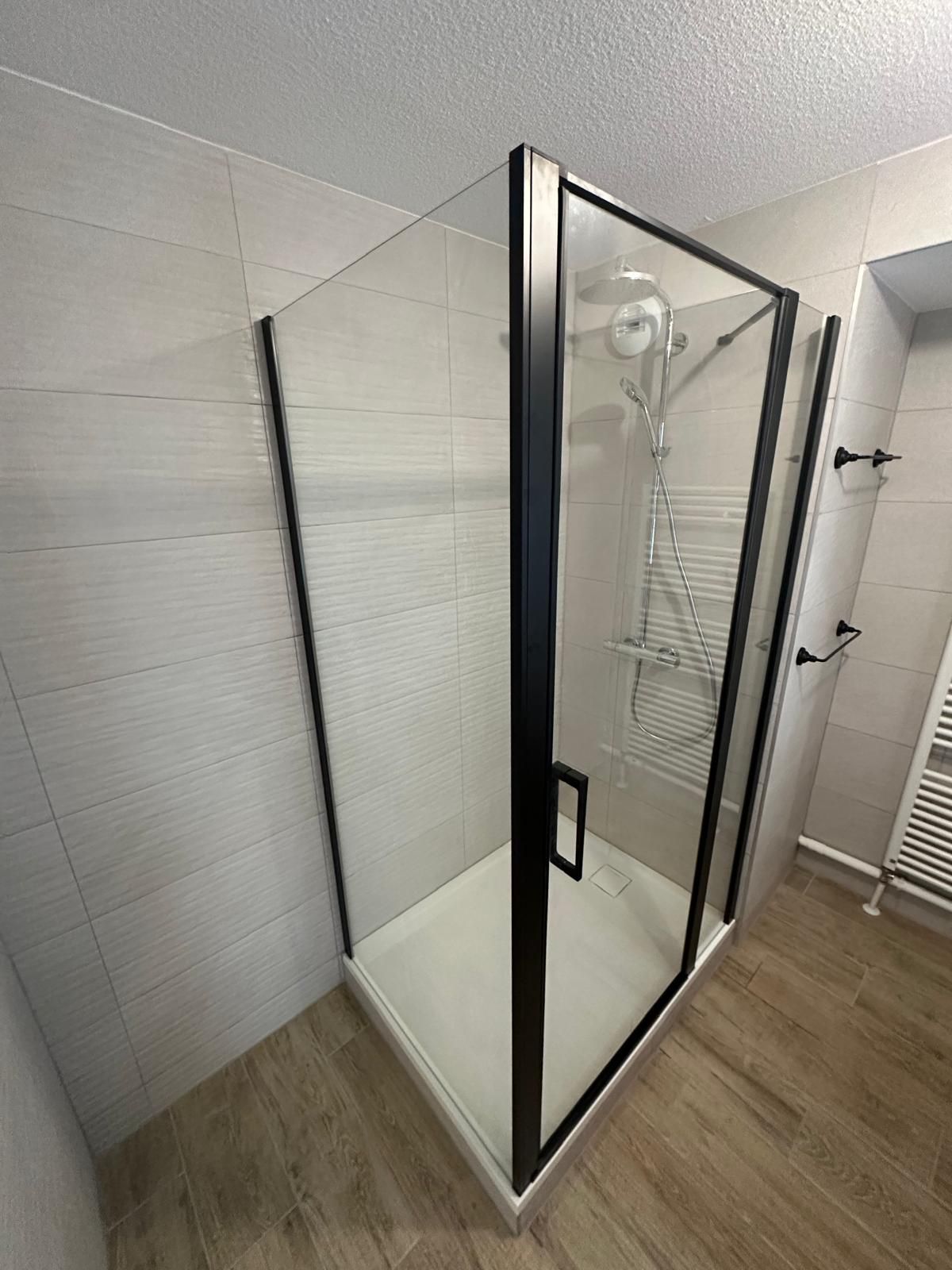 Cabine de douche en verre à cadre noir dans une salle de bains aux murs carrelés beiges et au socle blanc.