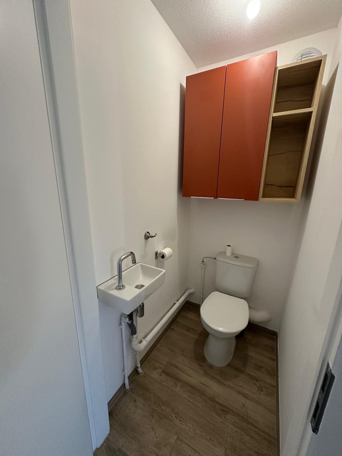 Petite salle de bains avec toilettes, lavabo et armoire de rangement rouge. Sol clair imitation bois et murs blancs.
