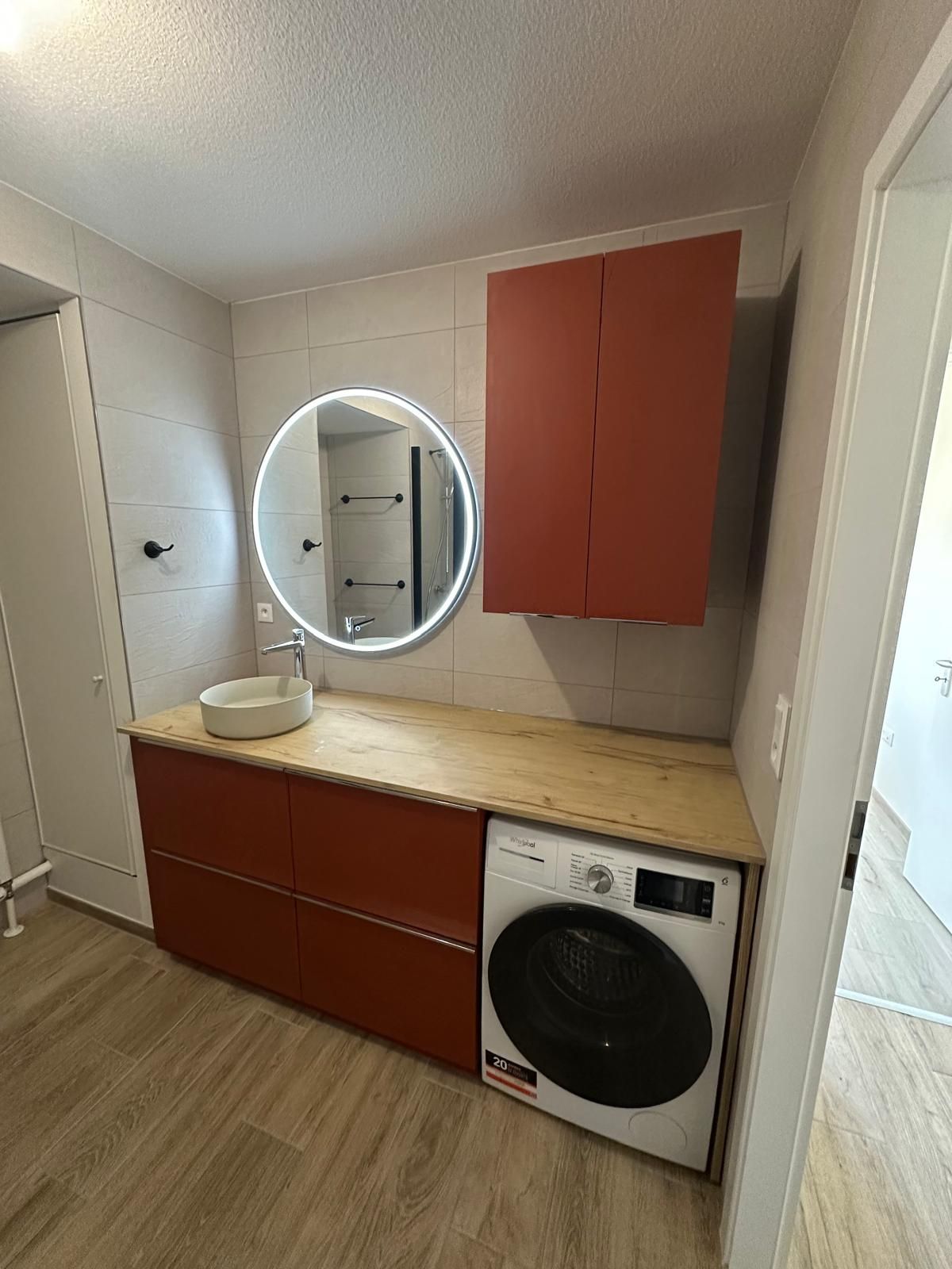 Salle de bains avec un meuble rouge, une machine à laver, un miroir rond et un lavabo.