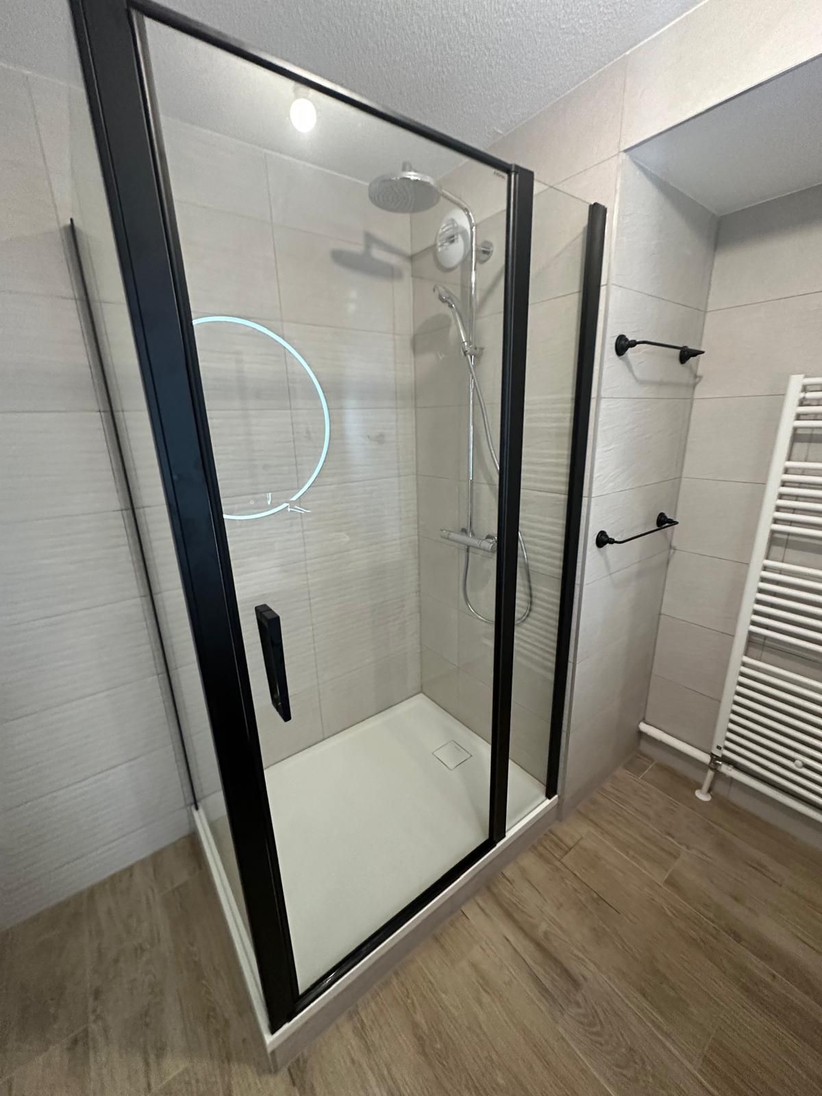 Douche en verre à cadre noir dans une salle de bains moderne avec carrelage clair, base blanche et sol imitation bois.