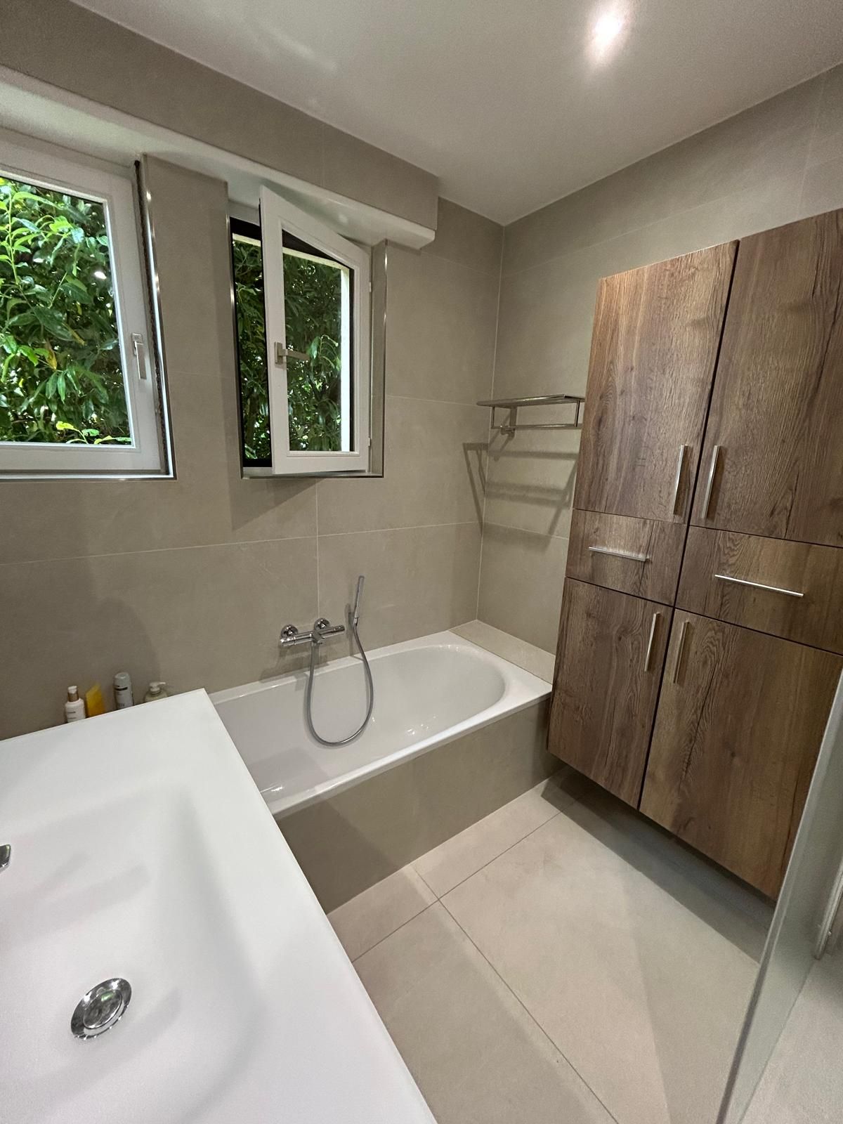 Salle de bains avec baignoire blanche, lavabo et carrelage aux tons neutres. Meuble en bois. Fenêtre ouverte donnant sur la verdure.