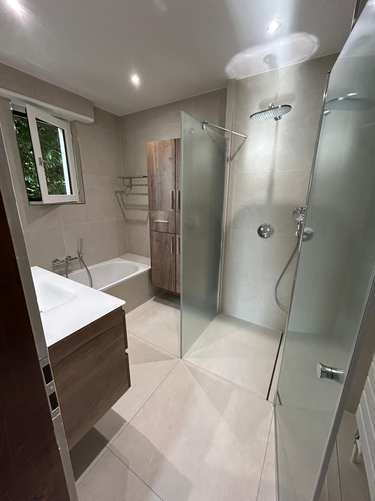 Salle de bains moderne avec douche, baignoire, meuble-lavabo et armoire en bois. Carrelage clair et verre dépoli.