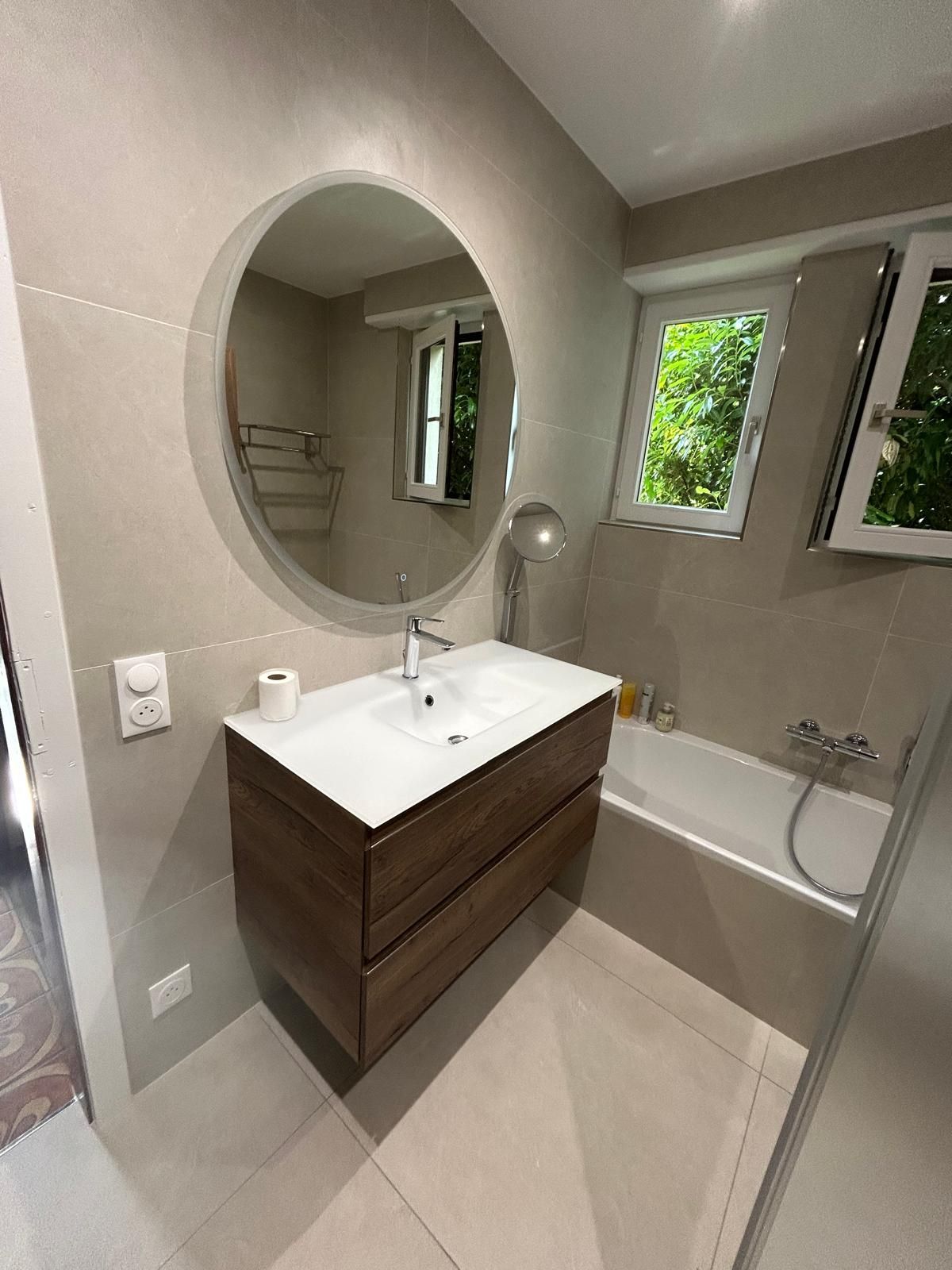 Salle de bains avec meuble-lavabo marron, miroir ovale, baignoire et fenêtre donnant sur la verdure.