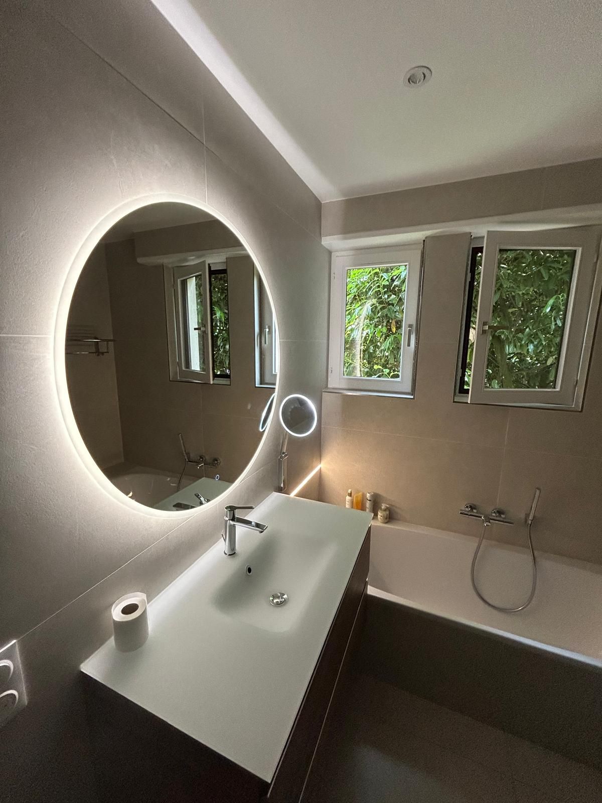 Salle de bains moderne avec un grand miroir circulaire éclairé, un lavabo rectangulaire et une baignoire ; deux fenêtres offrent une vue sur la verdure.