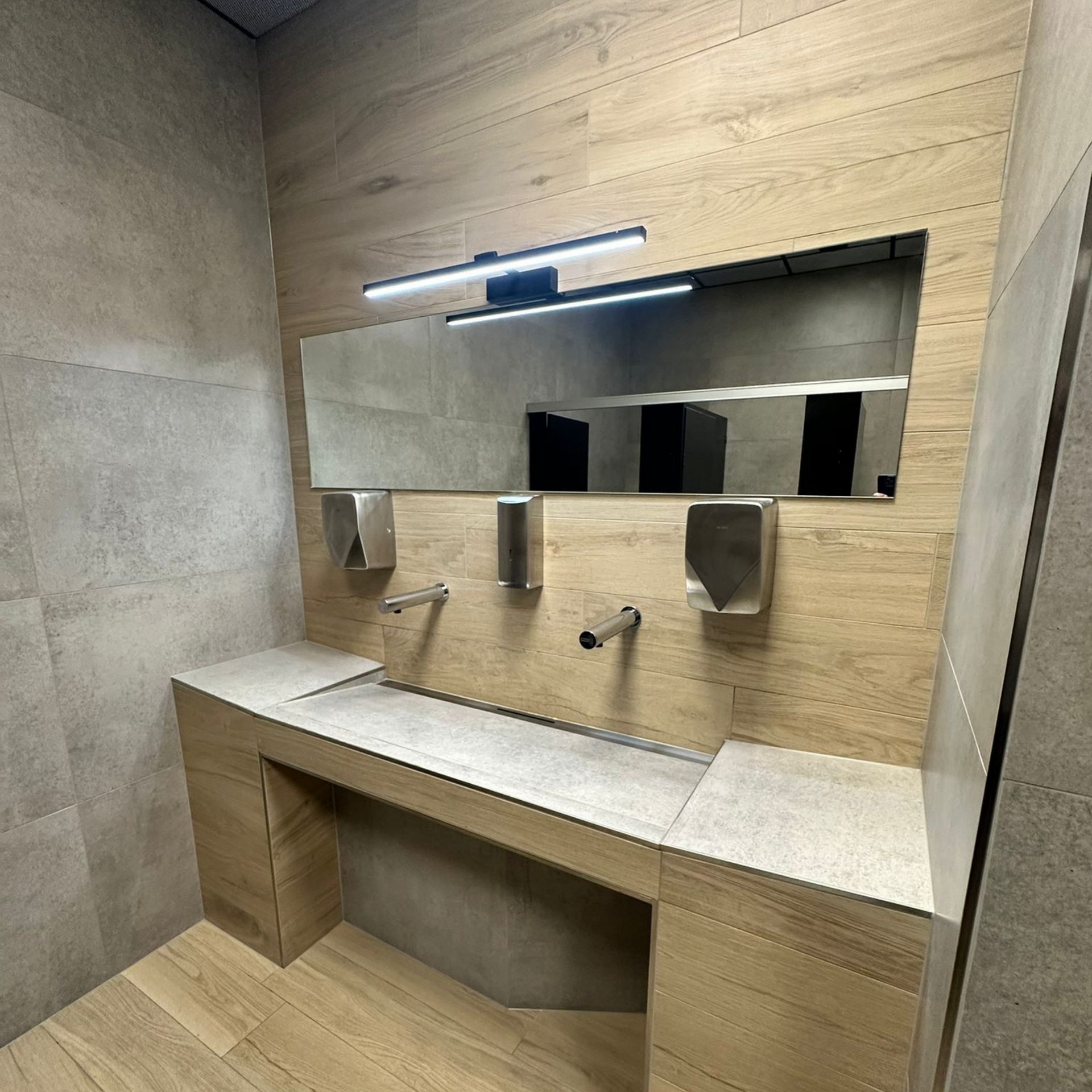 Lavabo de salle de bain avec miroir, distributeurs de savon et distributeurs d'essuie-mains en papier ; tons bois et gris.