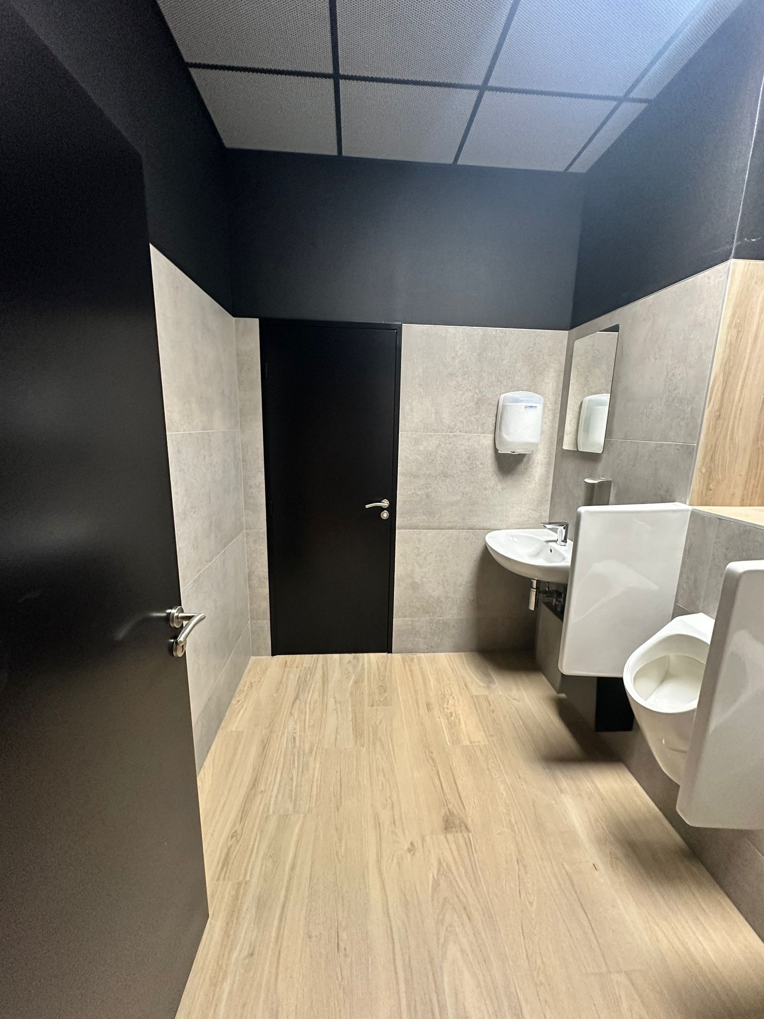 Intérieur de toilettes publiques avec parquet clair, murs gris pierre, porte noire et robinetterie blanche.