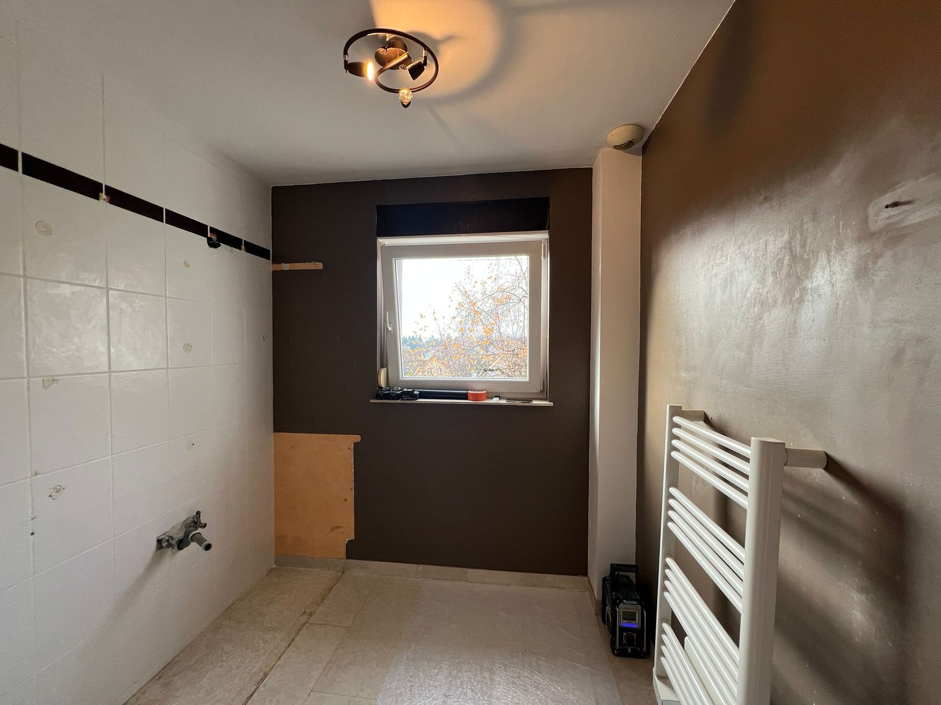 Salle de bains avec mur partiellement carrelé, fenêtre et murs et sol non finis.