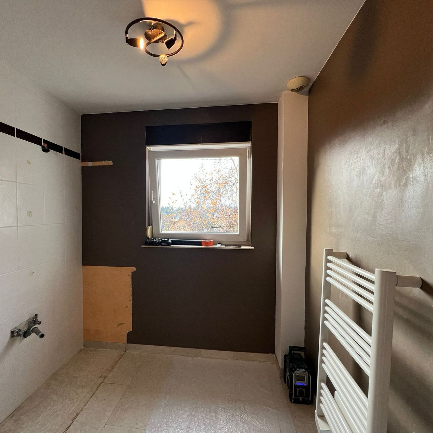 Salle de bains aux murs marron, carrelage blanc et fenêtre. Un radiateur blanc et un luminaire sont visibles.