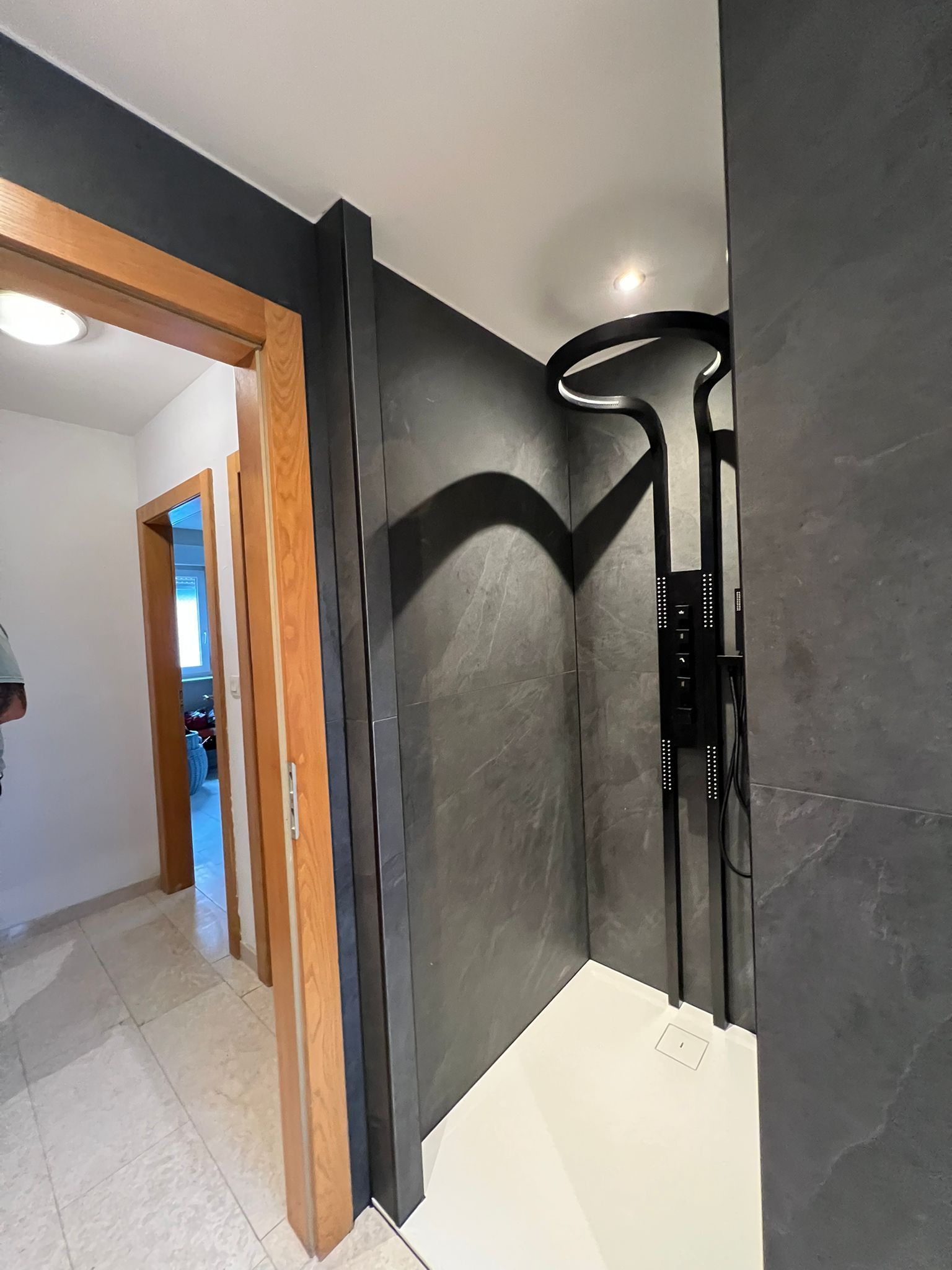 Couloir avec douche et porte. Paroi de douche grise et robinetterie noire. Encadrements de porte en bois.