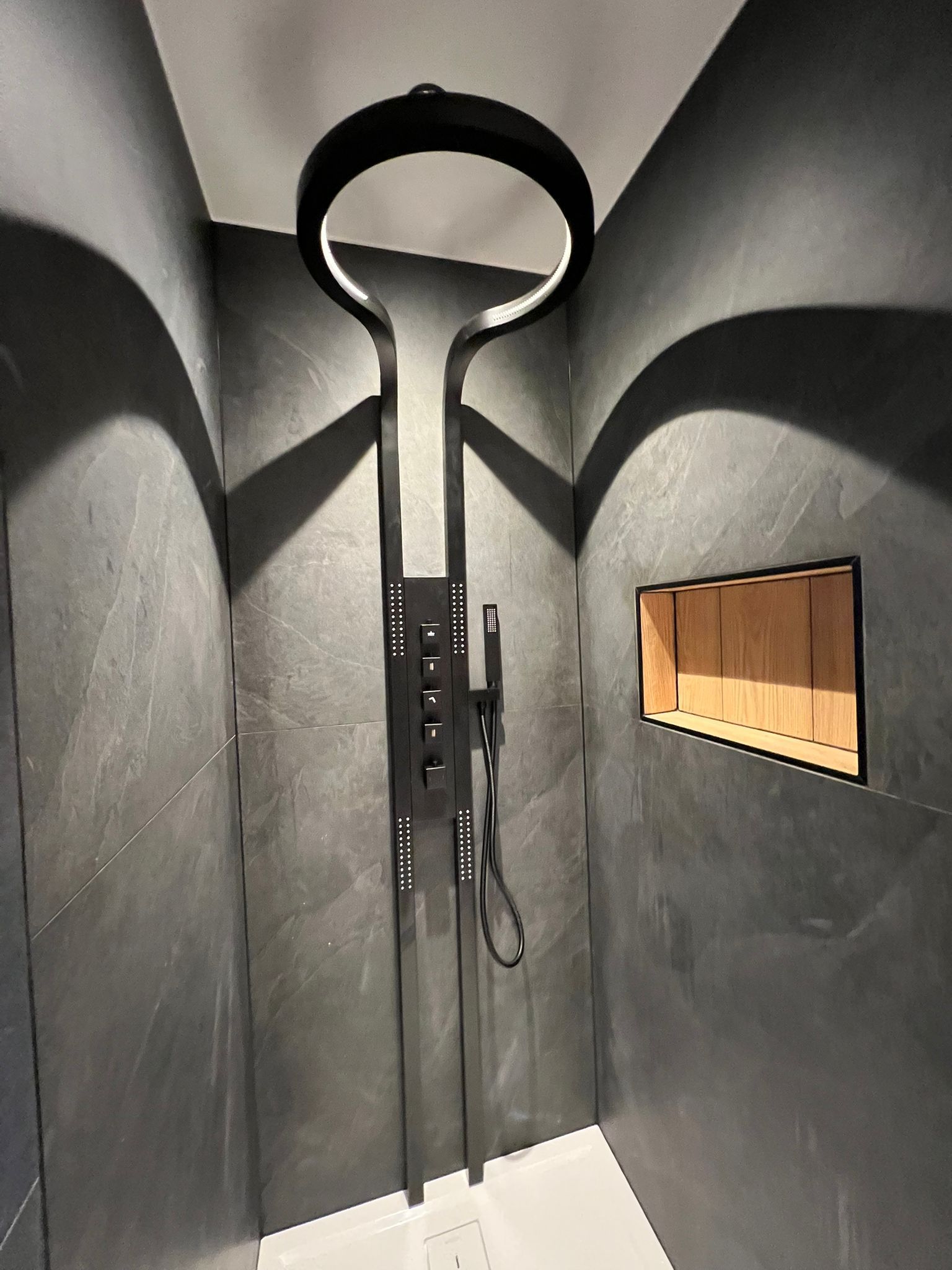 Douche moderne avec robinetterie noire et murs carrelés gris ; pommeau de douche arqué et étagère intégrée.