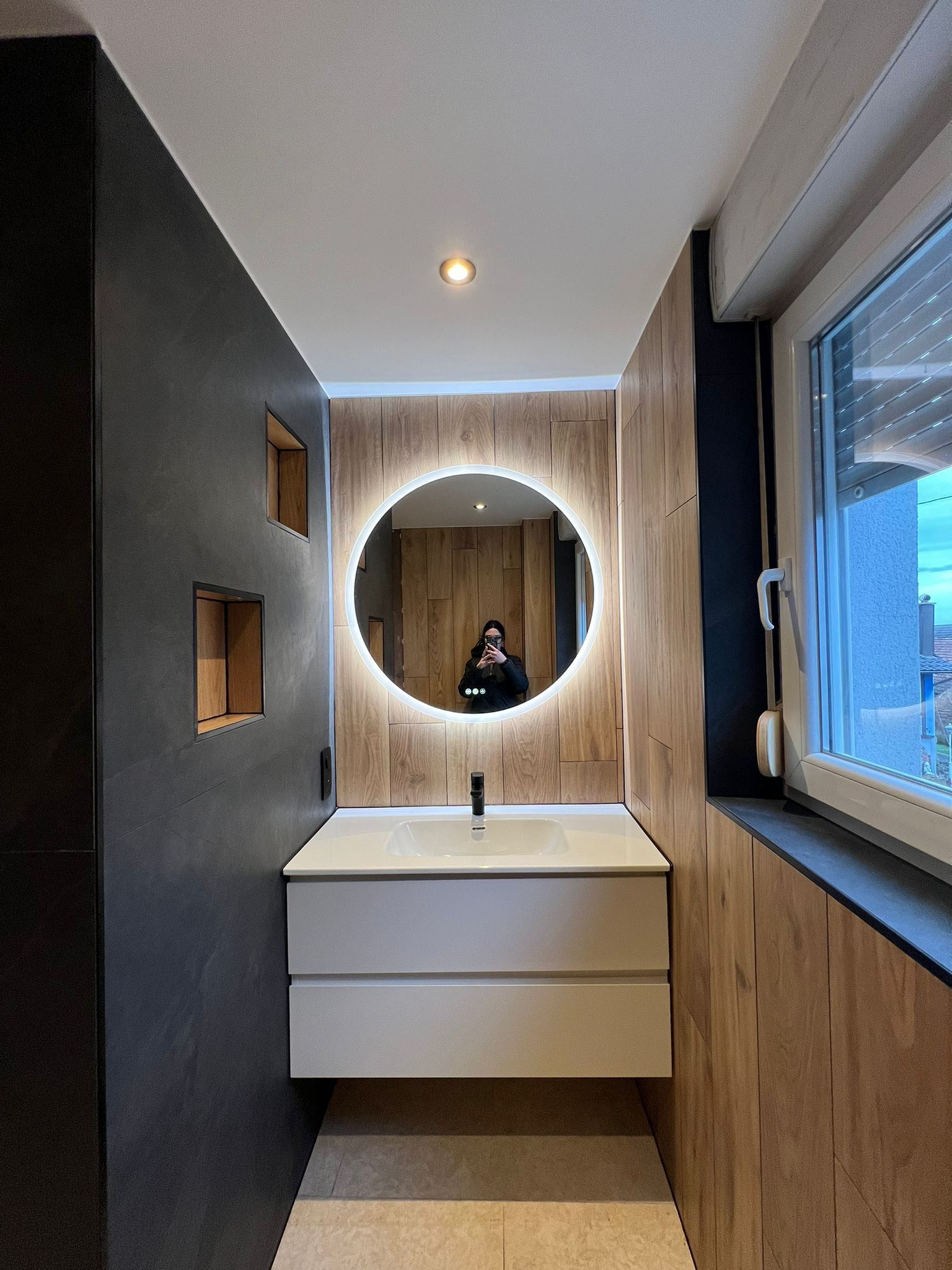 Salle de bains avec meuble-lavabo blanc, miroir rond avec éclairage et lambris aux murs.