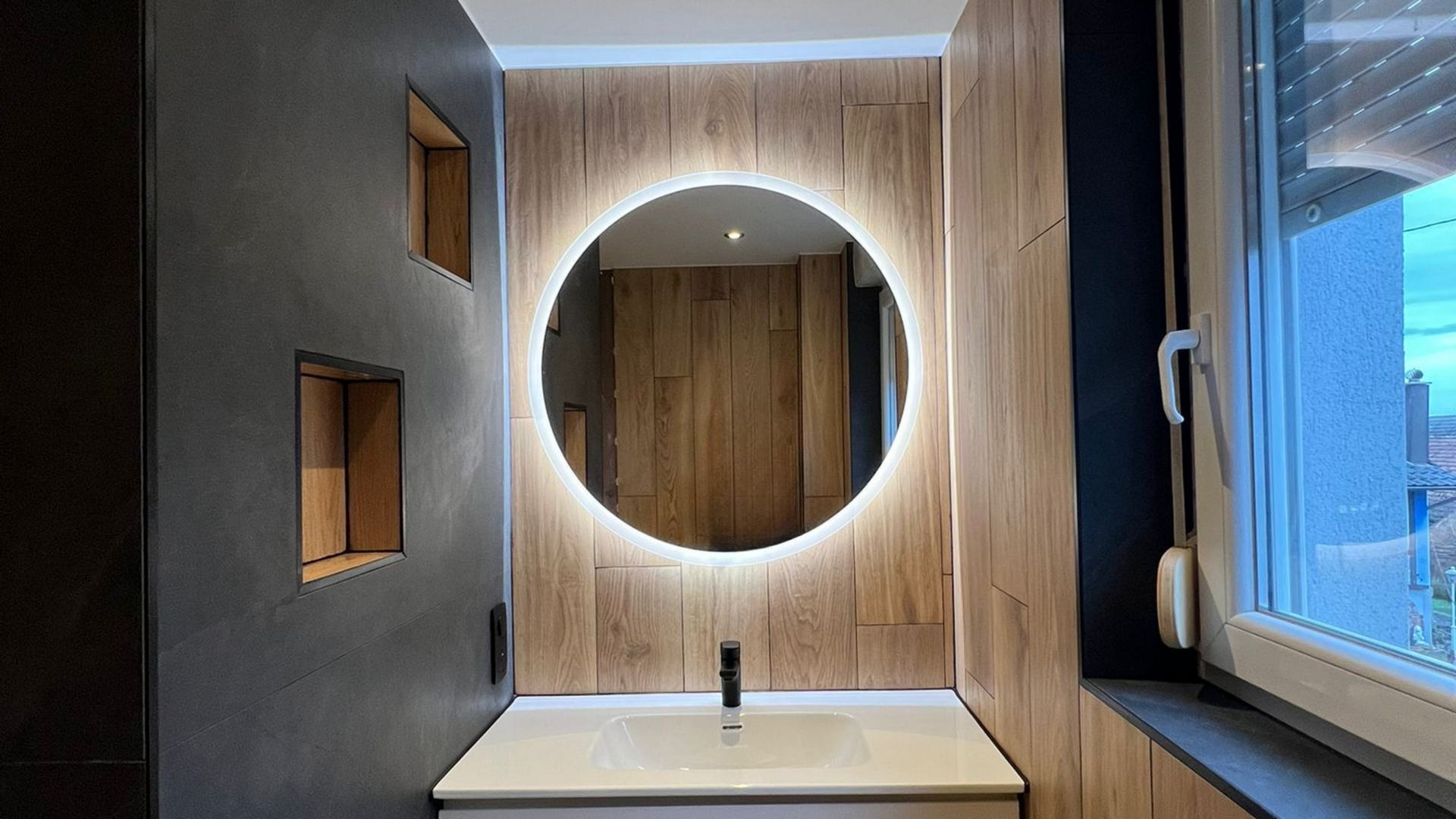 Salle de bains avec un miroir rond entouré de lumières LED, des lambris en bois et un lavabo blanc.