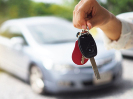 Main tenant des clés de voiture avec un porte-clés noir et une étiquette rouge devant une voiture argentée floue