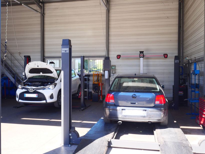 Deux voitures dans un garage, une blanche avec capot ouvert et une bleue sur un pont élévateur