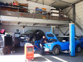 Intérieur d'un garage automobile : un mécanicien travaille sur un SUV bleu à côté d'une voiture noire sur un pont élévateur.
