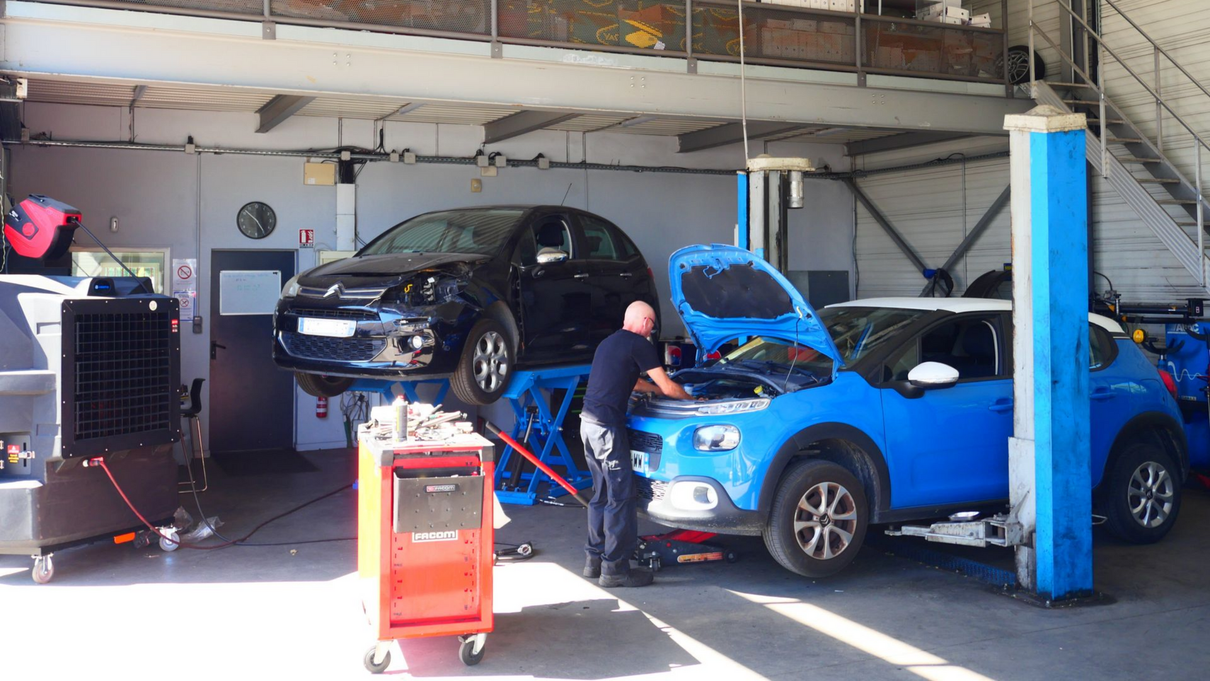 Un mécanicien travaille sur un SUV bleu dans un garage, une voiture noire se trouvant sur un pont élévateur derrière.
