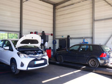 Deux voitures garées dans un garage, une voiture blanche avec capot ouvert et une voiture noire à côté.