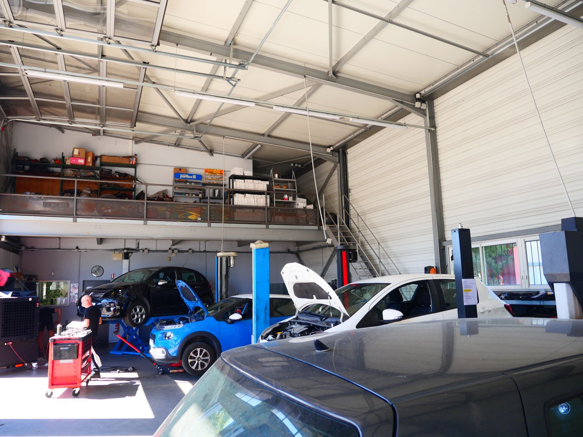 Garage automobile avec deux voitures bleues, l'une sur un pont élévateur et l'autre capot ouvert, et une voiture sombre au premier plan.
