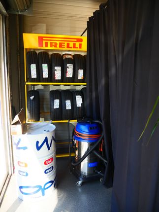 Zone de stockage de pneus Pirelli avec des pneus noirs empilés, un fût bleu et un conteneur blanc dans un hangar étroit