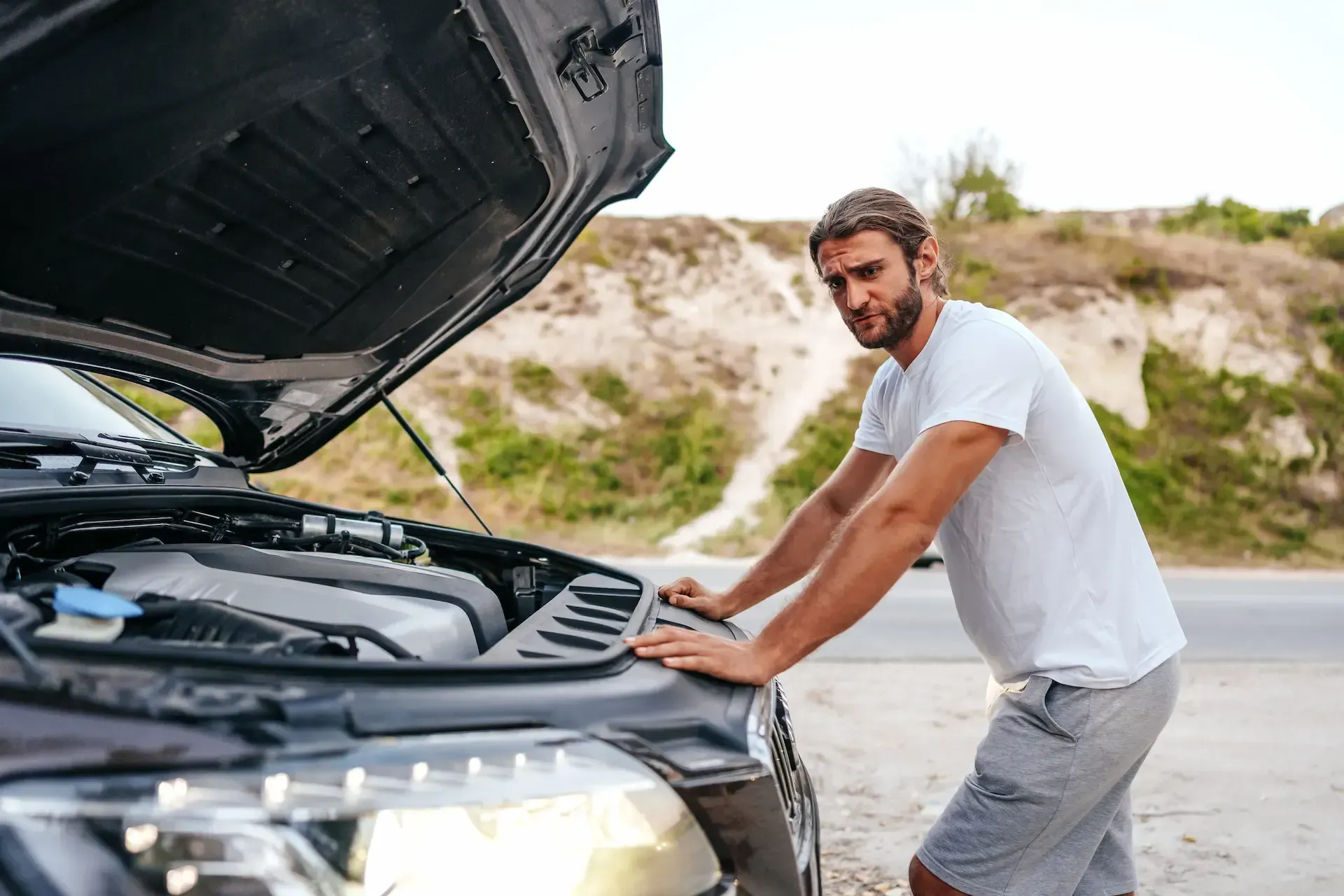 Hombre mirando el motor de un coche con el capó abierto, carretera y colinas al fondo.