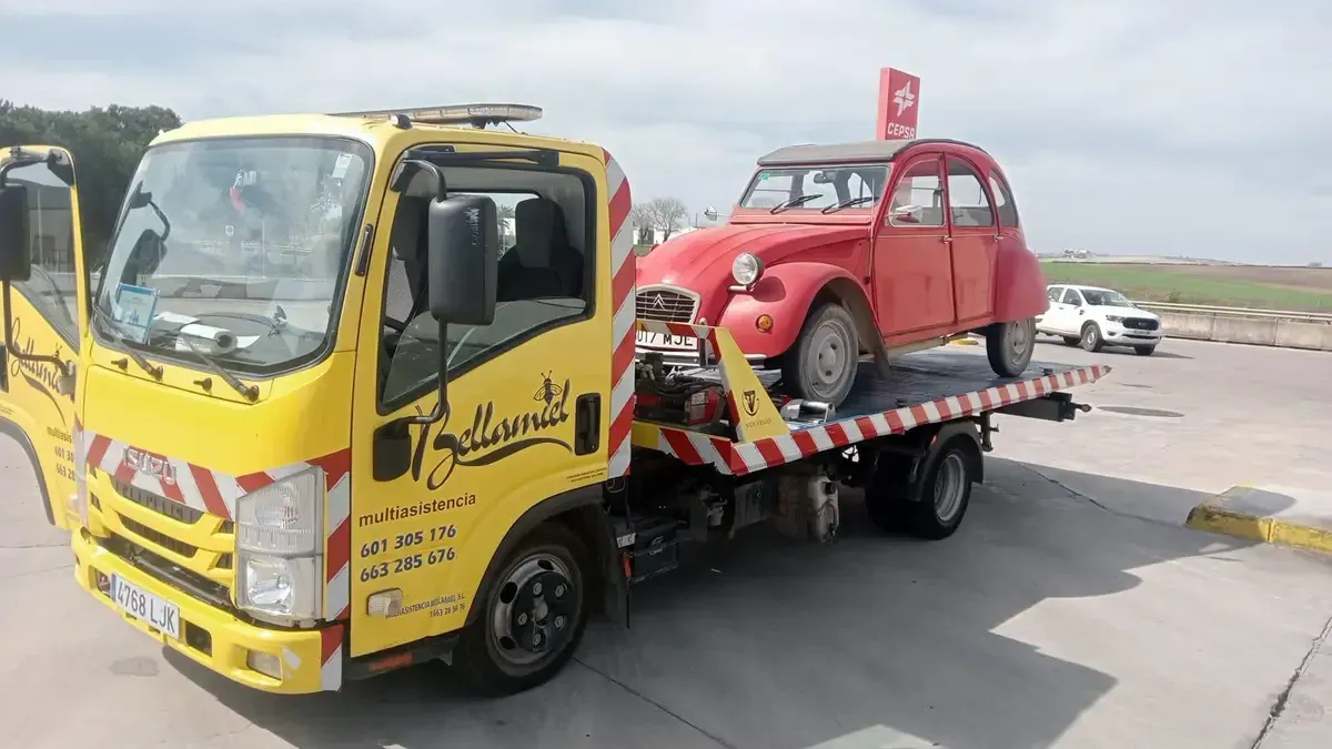 Grúa amarilla que transporta un coche Citroën 2CV rojo en una plataforma.