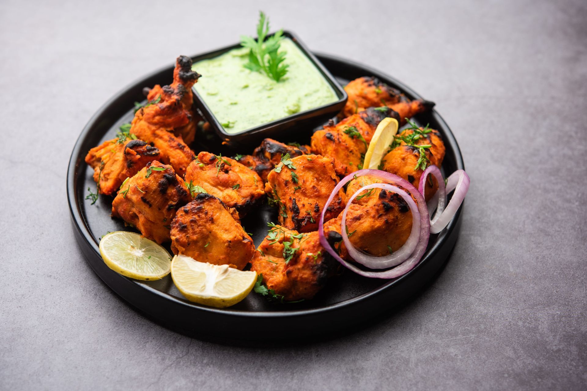 Pollo tandoori a la parrilla en un plato negro con rodajas de limón, aros de cebolla y chutney verde.