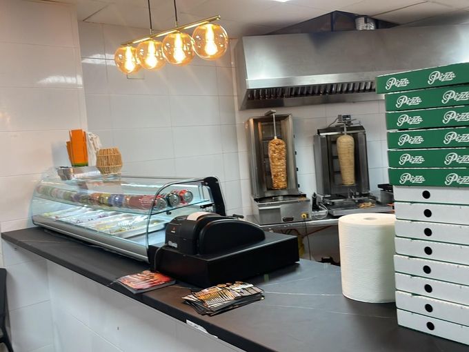 Mostrador de charcutería vacío con una vitrina de cristal, luces colgantes y cajas blancas apiladas en una tienda que parece una cocina.