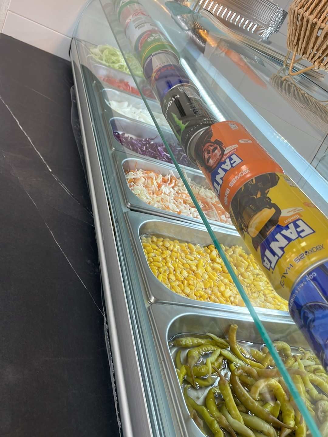 Barra de ensaladas con bandejas metálicas de verduras y encurtidos bajo una mampara de cristal.