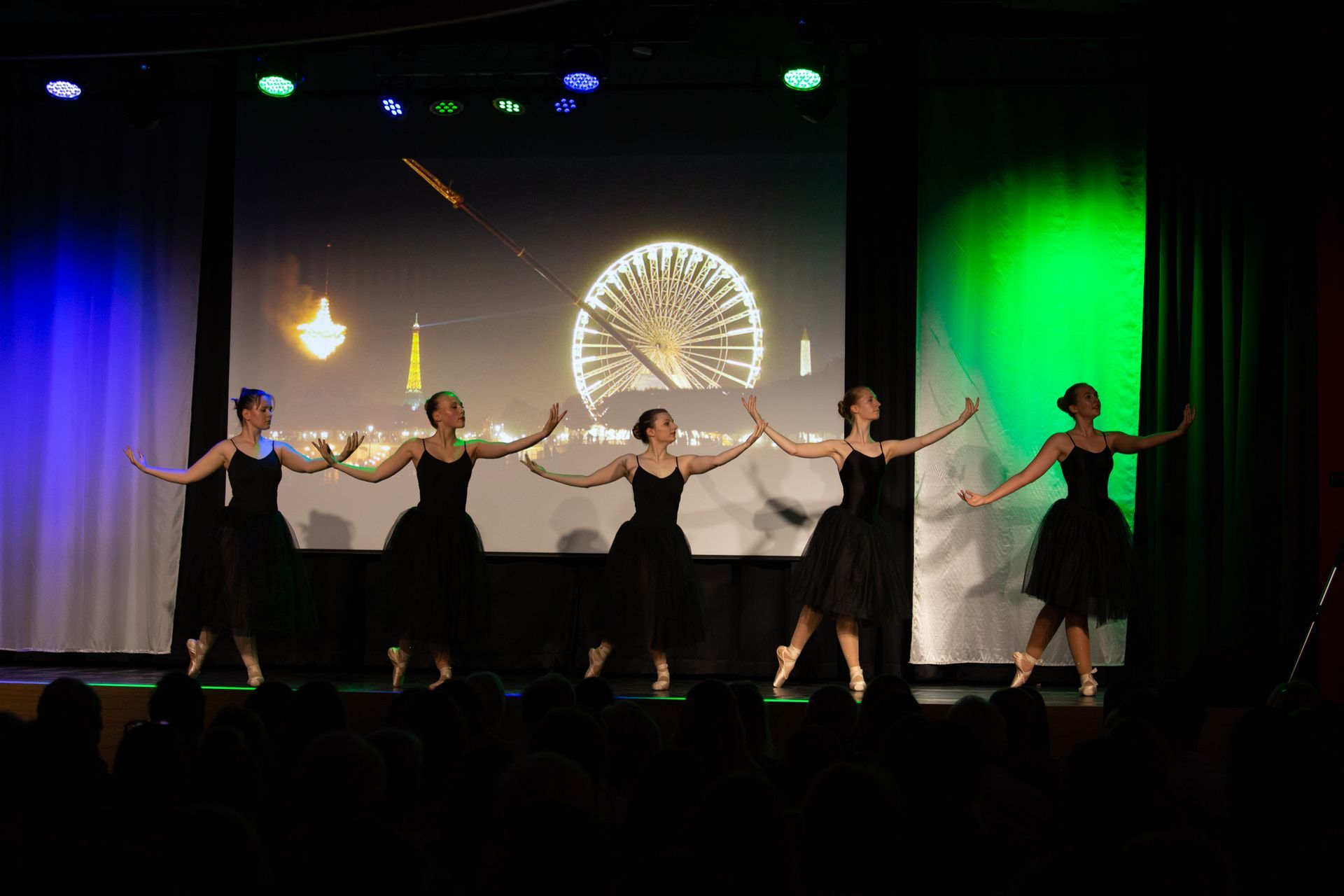 Tanzgruppe in schwarzen Ballett-Kleidern vor grün-blauer Lichtkulisse und einer Leinwand