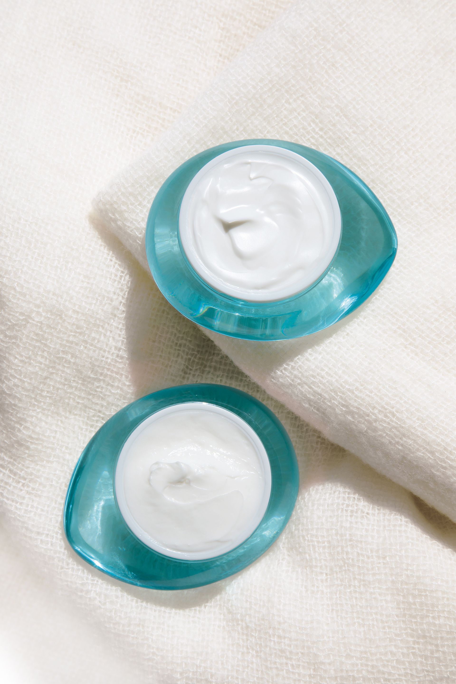 Deux pots de crème turquoise contenant de la crème blanche, posés sur un tissu beige doux.
