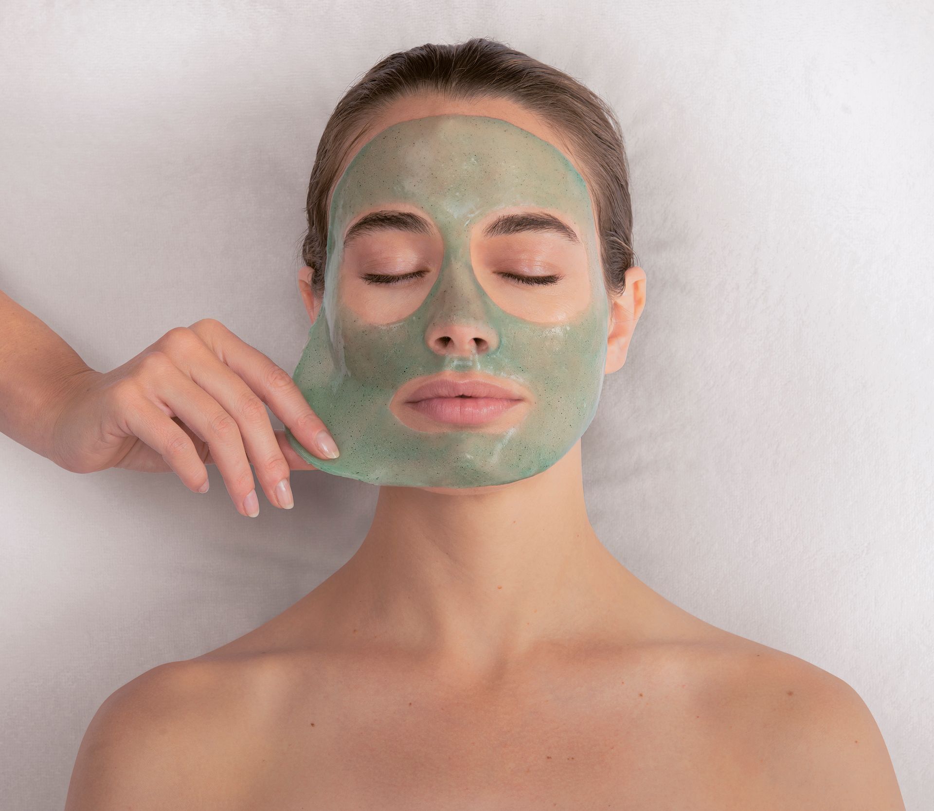 Femme portant un masque vert sur le visage, les yeux fermés, tandis qu’une main le retire sur fond blanc