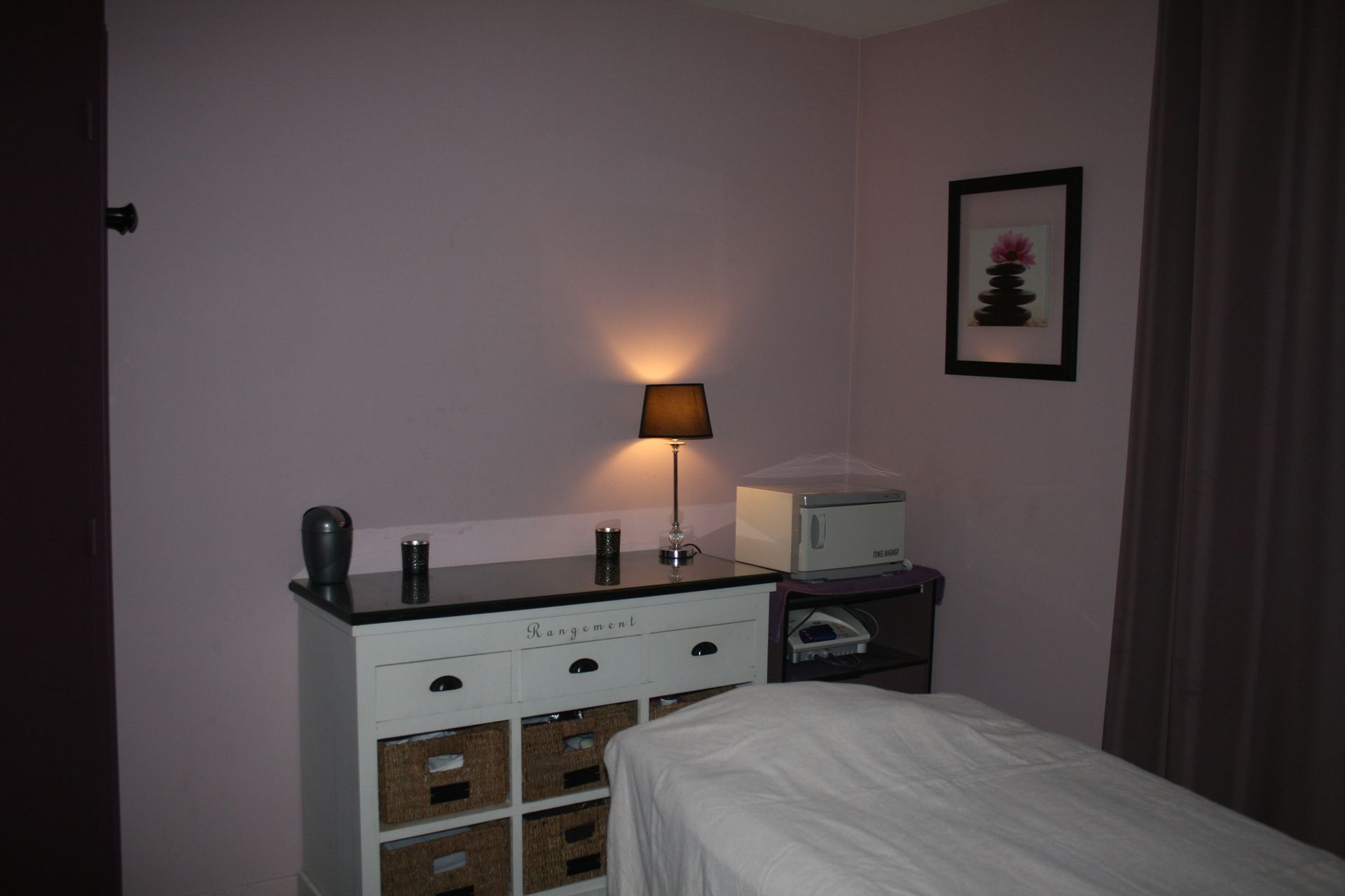 Chambre sombre avec un lit, une commode, une lampe et un tableau encadré.