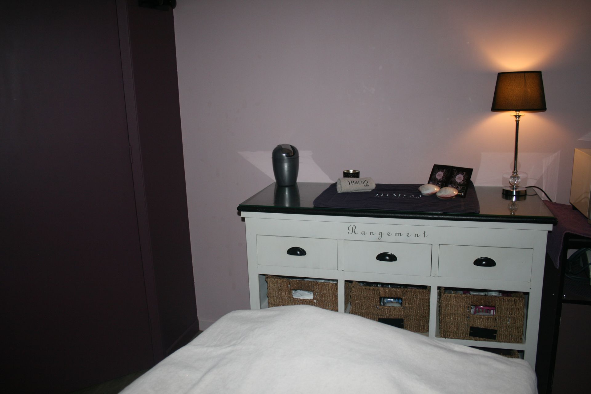 Chambre avec une commode blanche, une lampe allumée et des rideaux sombres à côté d’un lit au premier plan