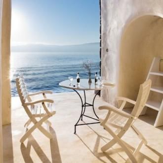 Deux chaises blanches et une table sur une terrasse ensoleillée au bord de la mer, avec vue sur une eau bleue et calme