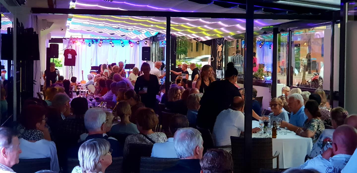 Ihmiset istuvat pöydissä ravintolan sisällä ja katsovat esitystä värikkäillä valoilla valaistulla lavalla.