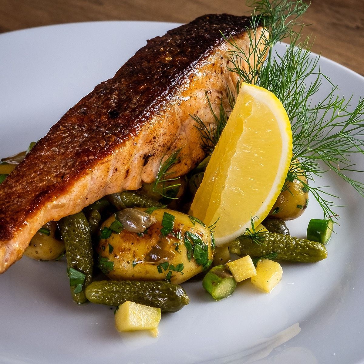 Paistettua lohifileetä sitruunan ja tillin oksan kera peruna- ja suolakurkkupedillä, tarjoiltuna valkoisella lautasella.