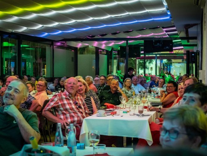 Ihmiset istuvat pöydissä huoneessa, jossa on värikkäät kattovalot ja joka näyttää tapahtumalta tai kokoontumiselta.