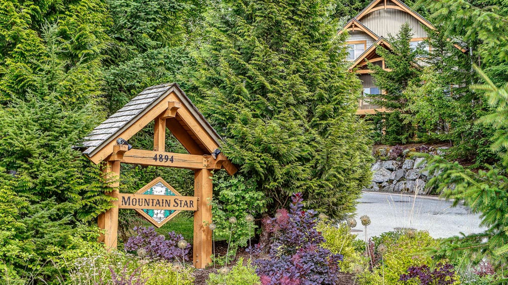 Wooden lodge sign amid lush green trees and purple flowers, with a cabin and stream in the background