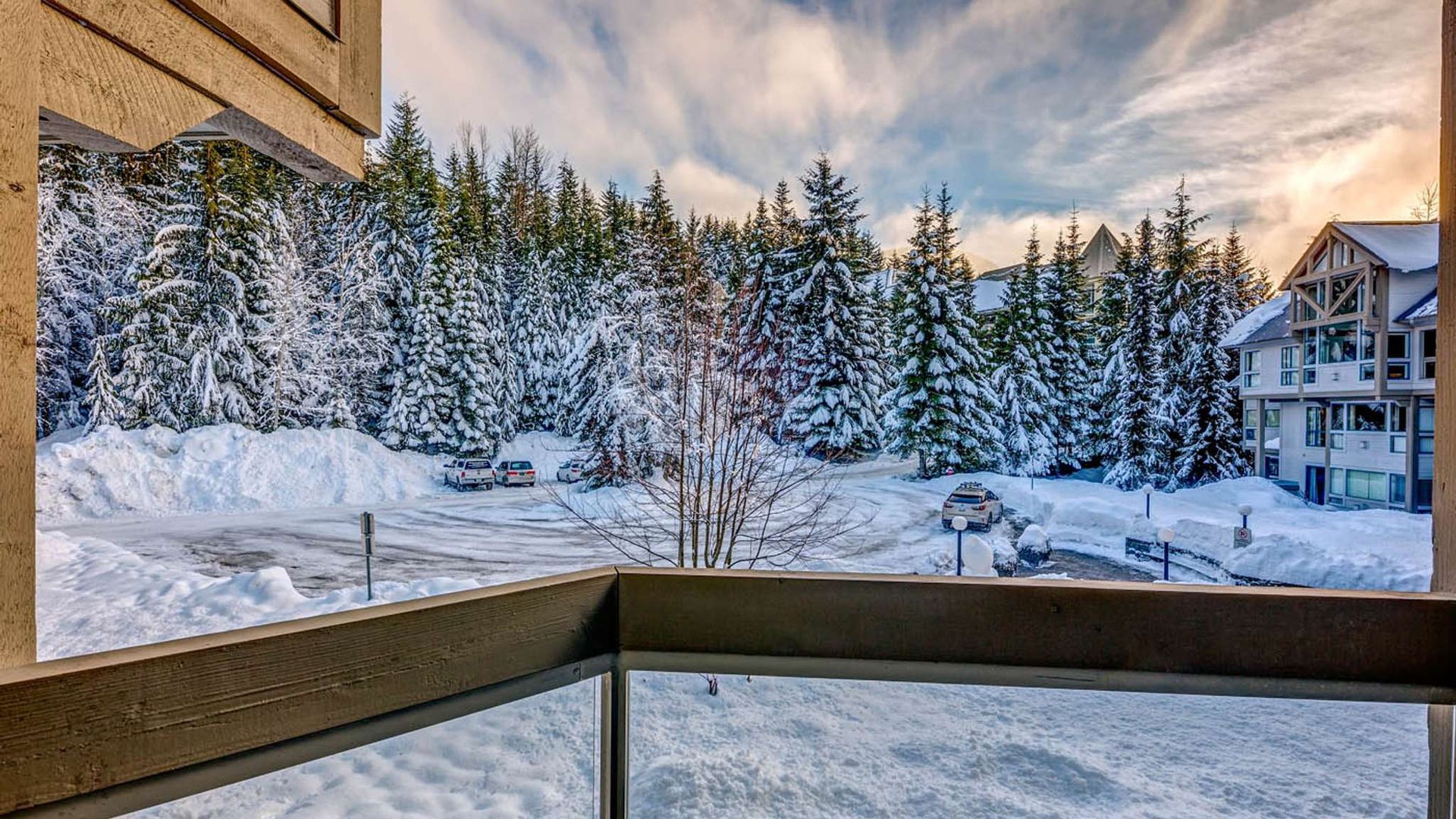 Snowy balcony view of pine trees and a condo at sunset