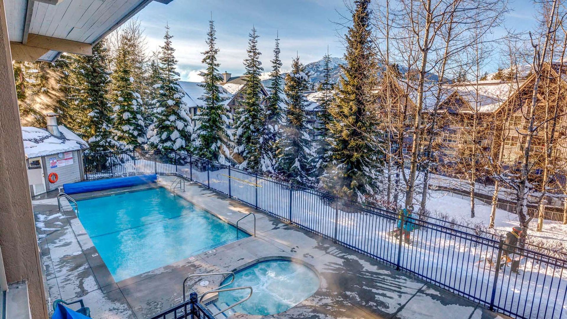 Outdoor pool and hot tub beside a snowy fence, with tall evergreen trees and mountains in the background